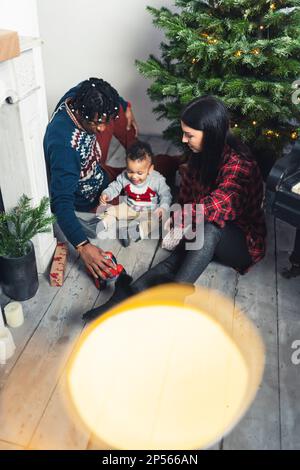 Multiethnische kleine Familie, die auf dem Boden vor dem Weihnachtsbaum sitzt und zusammen spielt, vertikaler Schuss. Hochwertiges Foto Stockfoto