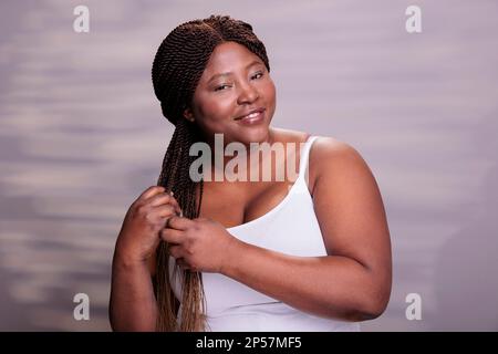 Kurvige glückliche Frau, die afrikanische Zöpfe frisiert und mit optimistischem Gesichtsausdruck in die Kamera schaut. Natürliche Schönheit gesunder Körper positive Frau posiert im Studio Stockfoto