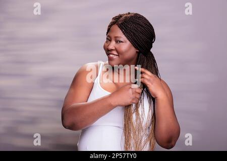 Wunderschönes lächelndes afroamerikanisches Mädchen, das lange braune Haare geflochten hat. Plus Größe Körper positiv hübsche Dame schaut in die Kamera und macht Zöpfe Haarstylistik, macht Haarpflege Routine Stockfoto