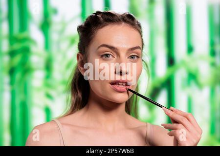 Attraktive Frau, die natürlichen Lipbalsam mit Pinsel aufträgt, sich mit organischen dekorativen Kosmetikporträts schminkt. Schönes junges Modell, das Lipgloss auf den Lippen trägt und in die Kamera schaut Stockfoto