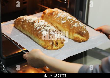 Zugeschnittenes Foto von Frauen Hände nehmen heißes, frisch gebackenes Brot aus einem elektrischen Ofen auf einem Metalltablett in der Küche zu Hause heraus. Rustikales hausgemachtes Brot, baki Stockfoto