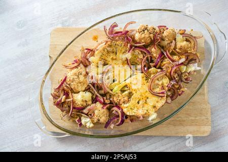 Gebackene Blumenkohl-Steaks mit würzigem Öl und roten Zwiebeln in einem Glasauflauf auf einem hölzernen Küchenbrett auf einem leichten Tisch, vegetarische Gemüsemehl, Spitze Stockfoto