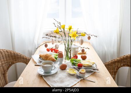 Frühstück für zwei mit Narzissen und Osterdekoration auf einem Holztisch am Fenster, Kopierbereich, ausgewählter Fokus, schmale Schärfentiefe Stockfoto