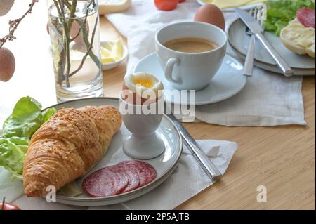 Frühstück oder Brunch mit Croissant, gekochtem Ei, Wurst, Käse und Kaffee auf leichten Servietten und einem Holztisch, ausgewählter Fokus, schmale Feldtiefe Stockfoto