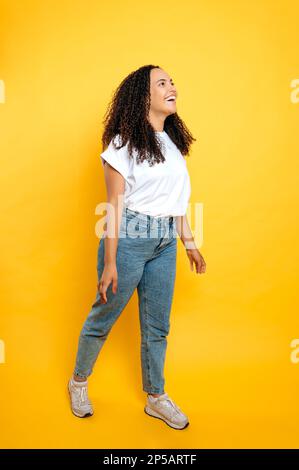 Langes Foto einer fröhlichen jungen brasilianischen oder lateinamerikanischen Frau mit lockigem Haar in lässiger Kleidung, die auf einem isolierten orangefarbenen Hintergrund steht, aufgeregt nach vorne blickt und glücklich lächelt Stockfoto