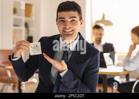 Ein junger Mann, der klebriges Papier mit Text zeigt... HAPPY IDIOTE's DAY in Office Stockfoto