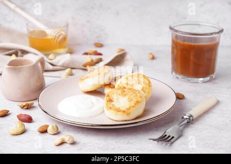 Teller mit leckeren Hüttenkäse-Pfannkuchen mit Sauerrahm, Karamellsirup und Nüssen auf hellem Hintergrund Stockfoto