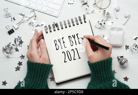 Frau schreibt "Bye Bye 2021" in einem Notizbuch auf einem weißen Holztisch, Draufsicht Stockfoto