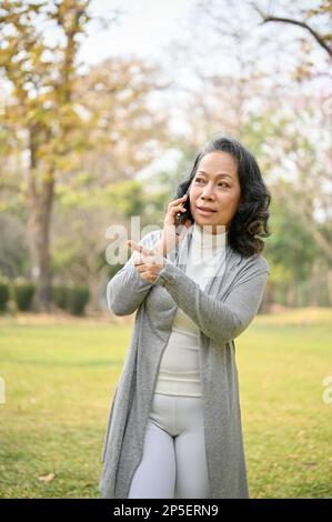 Porträt einer glücklichen 60-jährigen Asiatin in lässiger Kleidung, die mit jemandem telefoniert, während sie im grünen Park spaziert. Stockfoto