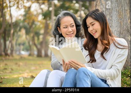 Glückliche und reizende asiatische Enkelin, die am Wochenende mit ihrer Großmutter unter dem Baum im wunderschönen grünen Park ein Buch liest. Stockfoto