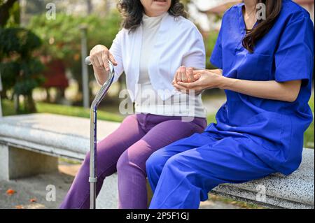 Zugeschnittenes Bild einer fürsorglichen asiatischen Krankenschwester oder Pflegekraft, die die Hand einer alten Patientin hält, spricht und Unterstützung gibt, während sie sich im Park entspannen Stockfoto