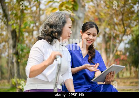 Eine glückliche und fürsorgliche junge asiatische Krankenschwester oder Pflegekraft zeigt einer alten Patientin etwas auf ihrem Tablet-Bildschirm, während sie spricht und sich auf einem Tisch ausruht Stockfoto