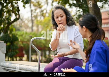 60s-jährige behinderte asiatische Frau nimmt eine Tablette, während sie sich mit ihrer Pflegekraft auf einer Bank in einem Park ausruht. Gesundheitskonzept Stockfoto