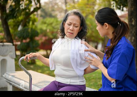 Unglückliche ältere behinderte asiatische Dame mit einem Gehstock, die auf einer Bank im Park sitzt, Rückenschmerzen hat und von ihrer Pflegekraft unterstützt wird. - Nein Stockfoto