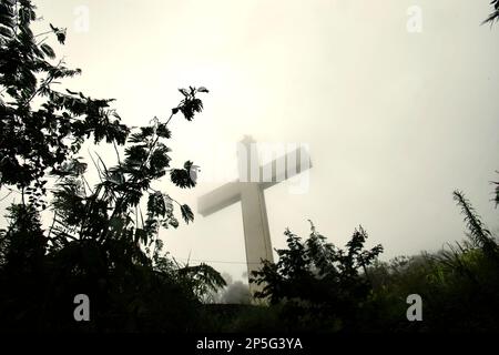 Ein hohes christliches Kreuz wird durch Nebel auf einem Hügel gesehen, der zu einem flachen Gelände des Hügels führt, wo Gotteshäuser aller anerkannten Religionen in Indonesien auf Bukit Kasih (Hügel der Liebe) gebaut werden, einem beliebten religiösen Reiseziel im Dorf Kanonang, West Kawangkoan, Minahasa, North Sulawesi, Indonesien. Gewidmet allen religiösen Gläubigen und Gläubigen, während sie die Geister der Liebe, des Friedens und der Toleranz fördern; Bukit Kasih wurde Anfang 2000 gegründet, als Adolf J. Sondakh Gouverneur der Provinz Nord-Sulawesi war. Es wurde als noble Friedensinitiative betrachtet. Stockfoto