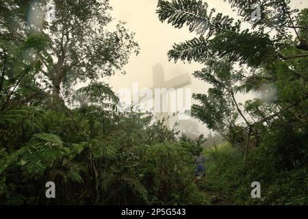 Ein hohes christliches Kreuz wird durch Nebel gesehen, im Vordergrund eines Dorfbewohners, der einen Pfad auf dem Bukit Kasih (Hügel der Liebe) hinuntergeht, einem beliebten religiösen Reiseziel im Dorf Kanonang, West Kawangkoan, Minahasa, North Sulawesi, Indonesien. Gewidmet allen religiösen Gläubigen und Gläubigen, während sie die Geister der Liebe, des Friedens und der Toleranz fördern; Bukit Kasih wurde Anfang 2000 gegründet, als Adolf J. Sondakh Gouverneur der Provinz Nord-Sulawesi war. Es wurde als noble Friedensinitiative betrachtet; eine Reaktion auf Konflikte, die auf Religion und ethnischer Zugehörigkeit beruhen, geschah vor und um... Stockfoto