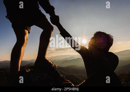 Silhouette eines Wanderers, der sich beim Sonnenuntergang gegenseitig beim Aufstieg auf einen Berg unterstützt. Menschen helfen und, Team arbeiten Konzept. Stockfoto