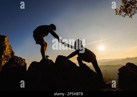 Silhouetten von zwei Menschen, die auf den Berg klettern und helfen. Hilfe- und Hilfekonzept. Stockfoto
