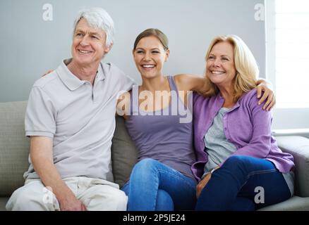 Es ist immer eine besondere Zeit, wenn unsere Tochter uns besucht. Ein glückliches älteres Paar, das sich zusammen mit seiner Tochter zu Hause auf dem Sofa entspannt. Stockfoto