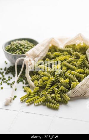 Grüne Mungbohnen-Fusilli-Pasta auf weißem Fliesenhintergrund. Beutel mit roher Pasta und Schüssel mit Mungbohne. Glutenfreie Nudeln. Stockfoto