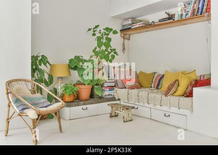 Ein Wohnzimmer mit vielen Pflanzen und Büchern auf den Regalen vor der Couch ist voller farbenfroher Kissen Stockfoto