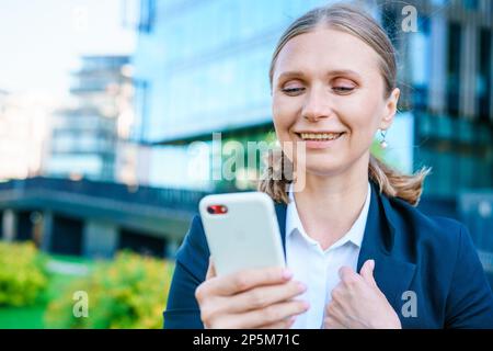 Erfolgreiche Bankerin, die im Freien ein Smartphone nutzt, während sie in der Nähe des Bürohintergrunds in Geschäftskleidung steht, junge Frau, die mit einem Mobilgerät in der Nähe eines Wolkenkratzers arbeitet Stockfoto