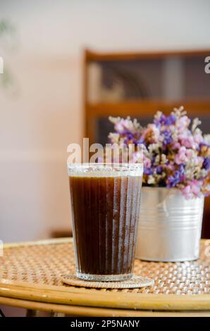 Ice-americano-Kaffee in großem Glas auf einem Holztisch mit violettem Blumenvase-Hintergrund Stockfoto