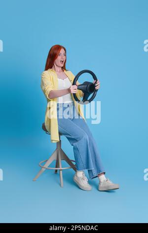Emotionale junge Frau auf einem Stuhl mit Lenkrad vor hellblauem Hintergrund Stockfoto
