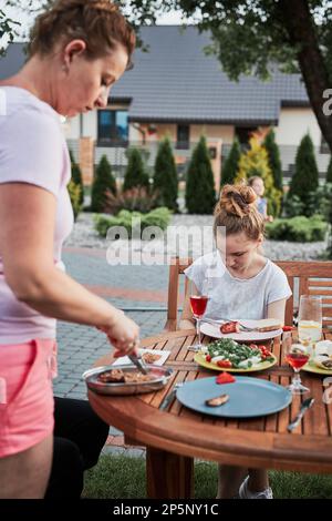 Familie mit einer Mahlzeit vom Grill während des Sommer Picknick im Freien Abendessen in einem Hausgarten. Nahaufnahme von Leuten, die an einem Tisch mit Essen und Geschirr sitzen Stockfoto