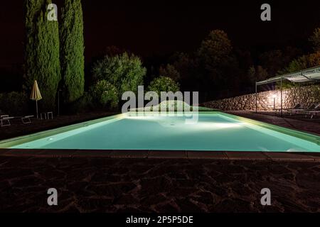 Beleuchteter Swimmingpool auf dem Hügel von Montemassi, umgeben von Zypressen und Oleandern in der Provinz Grosseto. Italien Stockfoto