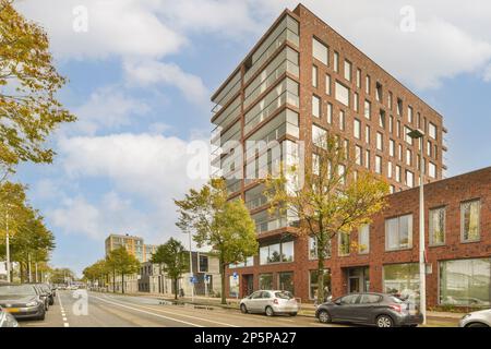 Eine Stadtstraße mit Autos, die am Straßenrand vor einem von Bäumen umgebenen Bürogebäude geparkt sind Stockfoto