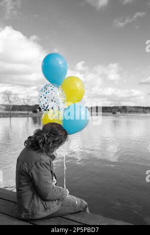 Schwarzweißes Porträt einer aufgewühlten, nicht erkennbaren Frau, die Ballons mit ukrainischer Flagge hält. Farbakzent auf Gelb und Blau. Stoppt den Krieg. Furcht A Stockfoto