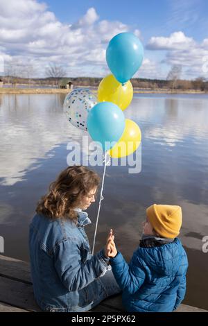 Eine junge Frau und ein Kind sitzen am Ufer des Sees und halten gelbe und blaue Ballons in den Farben der ukrainischen Flagge. Familie zusammen, du Stockfoto