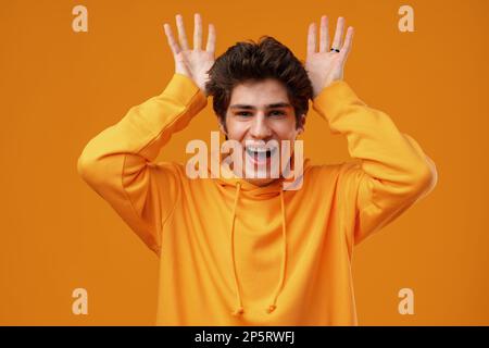 Ein Mann, der auf gelbem Hintergrund steht und eine lustige Geste macht, mit dem Finger über dem Kopf, als Stierhörner Stockfoto