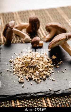 Seitenansicht von Trockenpulver aus Shiitake-Pilzen, Lentinula edodes. Lebensmittelzutat auf schwarzem Steinschneidbrett mit frischen Shiitake-Pilzen. Stockfoto