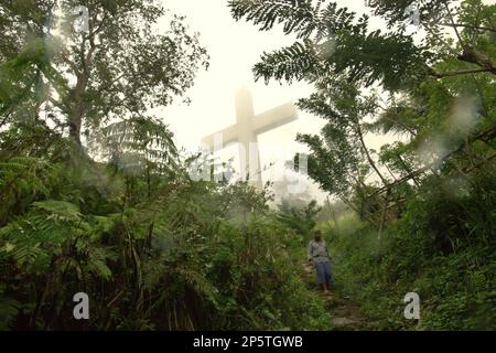 Ein hohes christliches Kreuz wird durch Nebel gesehen, im Vordergrund eines Dorfbewohners, der einen Pfad auf dem Bukit Kasih (Hügel der Liebe) hinuntergeht, einem beliebten religiösen Reiseziel im Dorf Kanonang, West Kawangkoan, Minahasa, North Sulawesi, Indonesien. Gewidmet allen religiösen Gläubigen und Gläubigen, während sie die Geister der Liebe, des Friedens und der Toleranz fördern; Bukit Kasih wurde Anfang 2000 gegründet, als Adolf J. Sondakh Gouverneur der Provinz Nord-Sulawesi war. Es wurde als noble Friedensinitiative betrachtet. Stockfoto