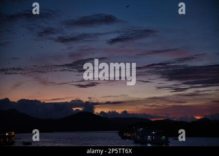 Romantische Aufnahme von leuchtenden Booten in der Mitte des Meeres bei einem intensiven roten, gelben orangefarbenen Sonnenuntergang am Himmel Hunderte von Fledermäusen. Stockfoto