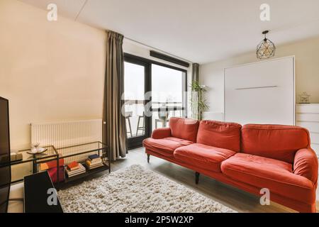 Ein Wohnzimmer mit roter Couch und weißem Teppich auf dem Boden vor dem Sofa ist ein offenes Fenster Stockfoto