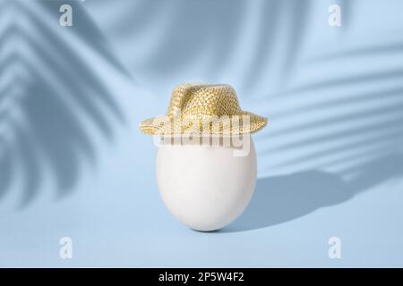 Einzelnes weißes Hühnerei in kleinem Sommerstrohhhut auf blauem Hintergrund mit Palmenblättern, Sommerferienkonzept, tropisches osterkonzept Stockfoto