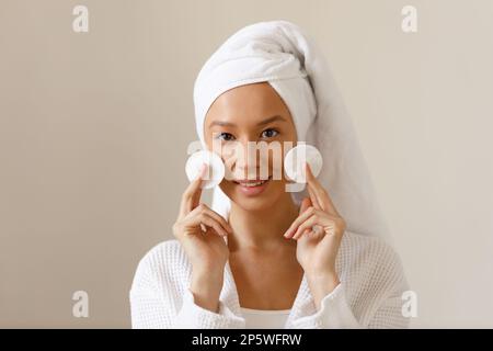 Eine junge Frau in weißem Bademantel und einem Handtuch am Kopf wischt die Haut ihres Gesichts mit Wattepads ab. Reinigung und gesunde Körperpflege nach dem Duschen. Nahaufnahme von Stockfoto