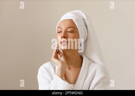 Eine junge Frau in weißem Bademantel und einem Handtuch am Kopf wischt die Haut ihres Gesichts mit Wattepads ab. Reinigung und gesunde Körperpflege nach dem Duschen. Nahaufnahme von Stockfoto