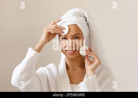 Eine junge Frau in weißem Bademantel und einem Handtuch am Kopf wischt die Haut ihres Gesichts mit Wattepads ab. Reinigung und gesunde Körperpflege nach dem Duschen. Nahaufnahme von Stockfoto