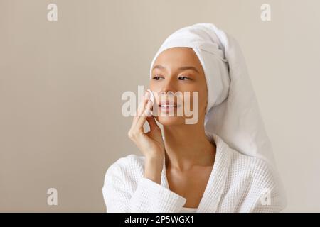 Eine junge Frau in weißem Bademantel und einem Handtuch am Kopf wischt die Haut ihres Gesichts mit Wattepads ab. Reinigung und gesunde Körperpflege nach dem Duschen. Nahaufnahme von Stockfoto