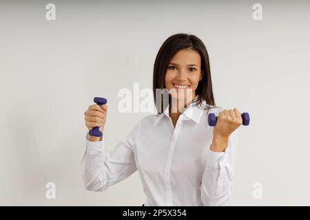 Eine attraktive Frau in einem weißen Hemd hält kleine weibliche Kurzhanteln. Porträt einer fröhlichen, lächelnden jungen Frau mit Sportausrüstung auf isoliertem weißem Hintergrund Stockfoto