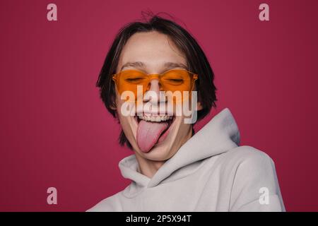 Porträt einer sorglosen Person in einer stilvollen Sonnenbrille, die aus der Zunge herausragt, isoliert auf pinkfarbenem Studiohintergrund. Jugendbegriff, Mode, Emotionen Stockfoto