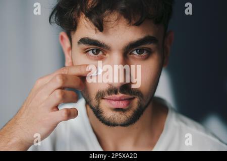 Ein weißer Mann, der in die Kamera schaut, die feuchtigkeitsspendende Creme auf die Wange aufträgt, beruhigt und weicht empfindliche Haut. Morgenhygiene. Stockfoto