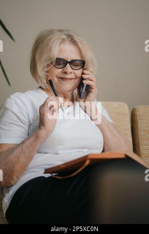 Porträt einer attraktiven, fröhlichen Seniorin, die auf Diwan sitzt, Kunden kommuniziert und in Notizblock schreibt und zu Hause an einem Projekt arbeitet. Smartphone Stockfoto