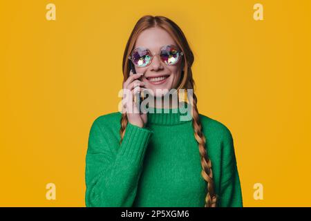 Foto einer attraktiven, positiv lächelnden jungen, rothaarigen Frau mit stilvollem grünem Pullover und Sonnenbrille, isoliert auf gelbem Hintergrund, die kommuniziert Stockfoto