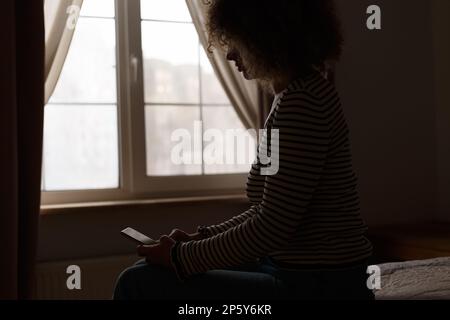 Silhoutte einer jungen Frau, die auf dem Bett im dunklen Schlafzimmer sitzt und eine Nachricht auf dem Smartphone tippt Stockfoto