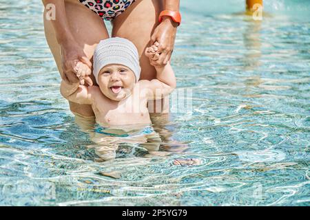 Die Mutter hält die Hände eines kleinen Jungen, der im Pool mit klarem blauem Wasser steht. Süßes Kind mit ausgestreckter Zunge, das in die Nahaufnahme der Kamera schaut Stockfoto